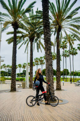 advertising photography of young woman standing with collapsible bike by a palm tree